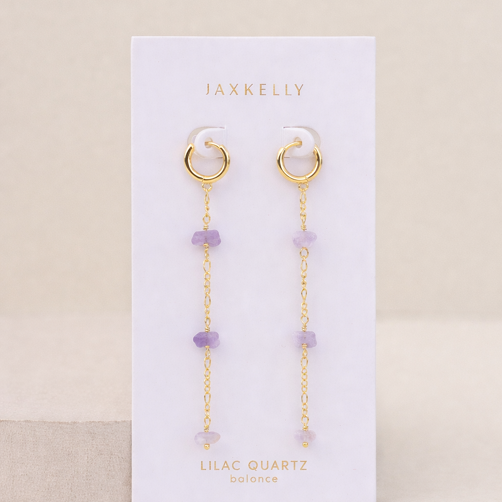 The Raw Gemstone Chain Drop Earring - Lilac Quartz features gold chains with lilac quartz beads, displayed on a white JaxKelly card labeled "Lilac Quartz balance." Tarnish-free and perfect for everyday elegance against a neutral beige background.