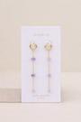 The Raw Gemstone Chain Drop Earring - Lilac Quartz features gold chains with lilac quartz beads, displayed on a white JaxKelly card labeled "Lilac Quartz balance." Tarnish-free and perfect for everyday elegance against a neutral beige background.