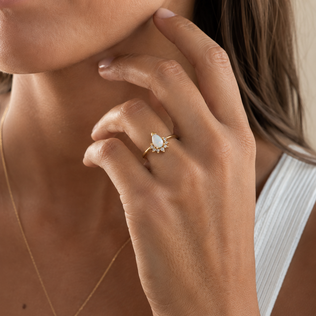 A woman touches her face while wearing the JaxKelly Opal Burst Ring—a gold plated design with a teardrop-shaped white cubic zirconia and accent stones—paired with a gold necklace and a white textured top.