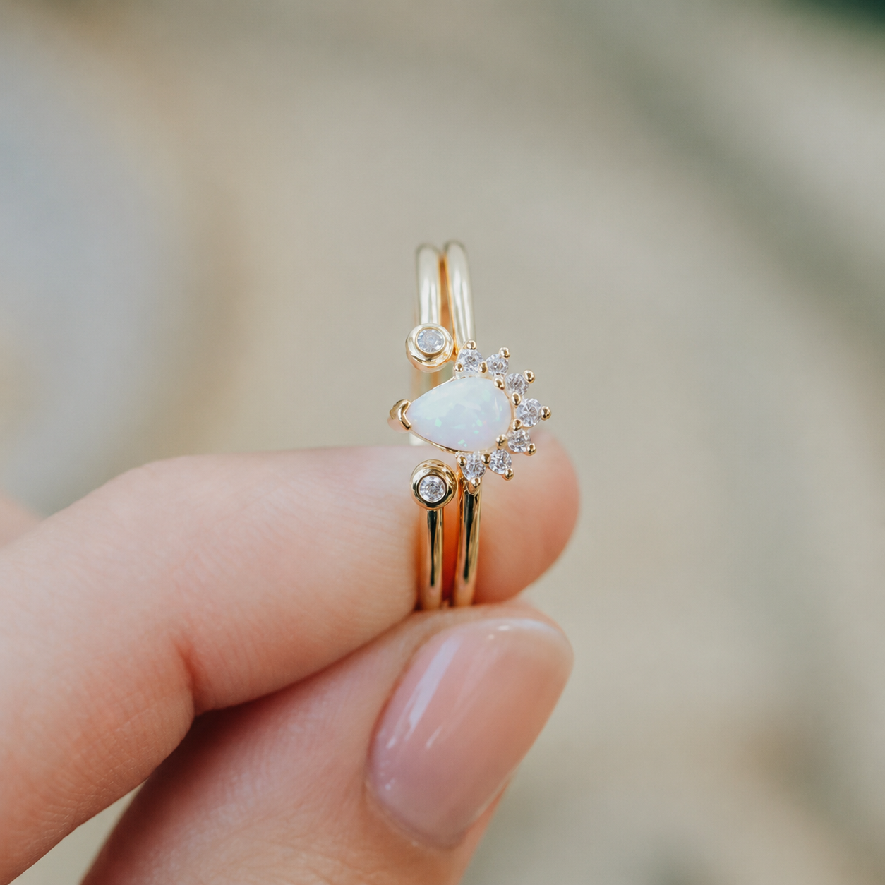 A hand holding the JaxKelly Opal Burst Ring, an 18K gold plated set with a lab-grown white opal teardrop, sparkling diamond accents, and a distinctive sunburst design.