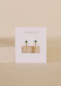 The JaxKelly Open Mini Hoop Earrings - Green Emerald feature 18K gold plating and small emerald CZ stones, displayed on a white JaxKelly card against a neutral beige background.