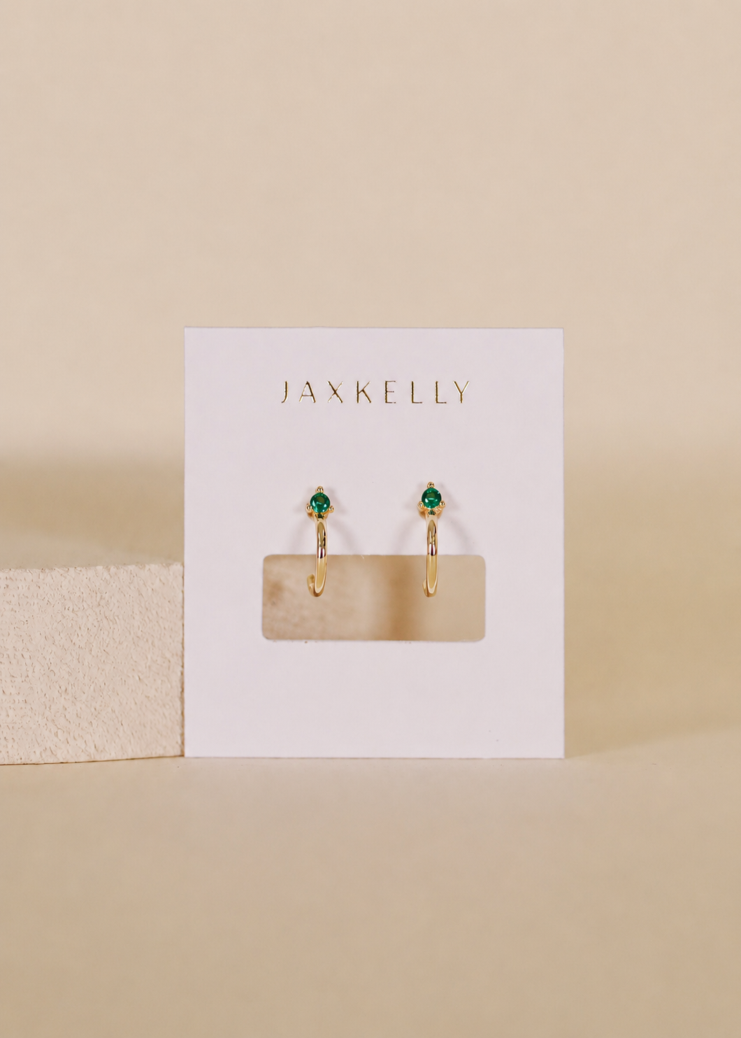 The JaxKelly Open Mini Hoop Earrings - Green Emerald feature 18K gold plating and small emerald CZ stones, displayed on a white JaxKelly card against a neutral beige background.