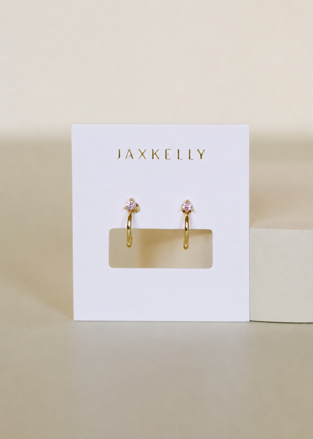 The JaxKelly Open Mini Hoop Earrings in Pink feature gold plating and pink stone accents, are hypoallergenic for sensitive ears, and come displayed on a white JaxKelly card against a neutral background.