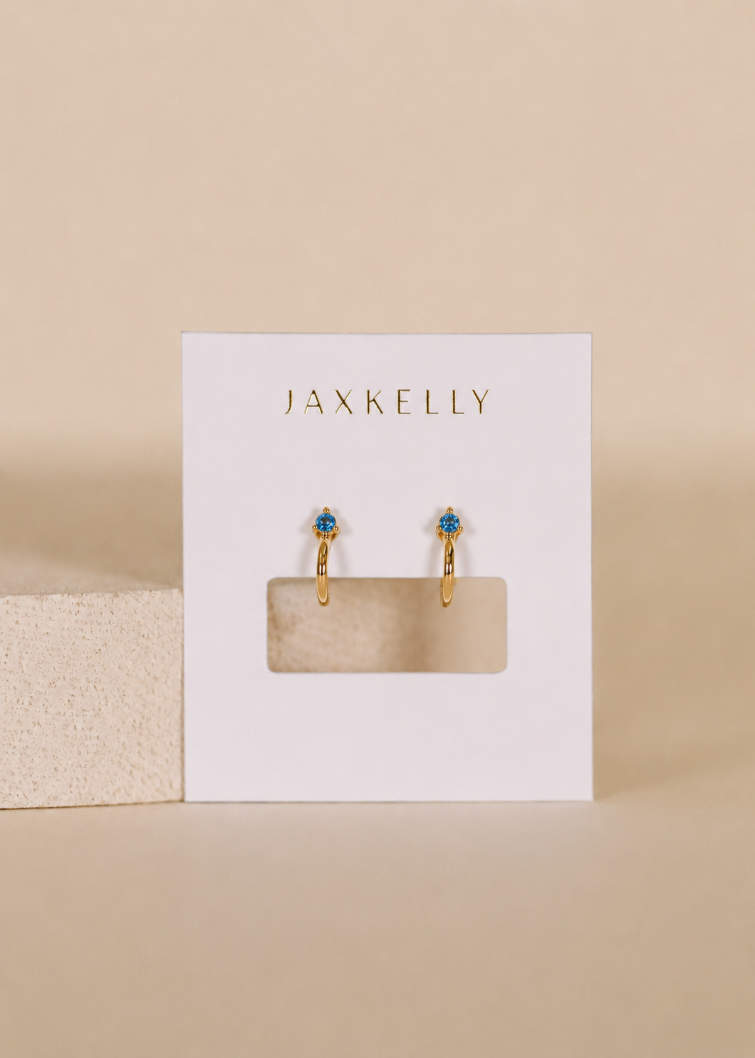 The Open Mini Hoop Earrings – Blue Sapphire by JaxKelly are shown on a white JaxKelly card against a neutral beige background.