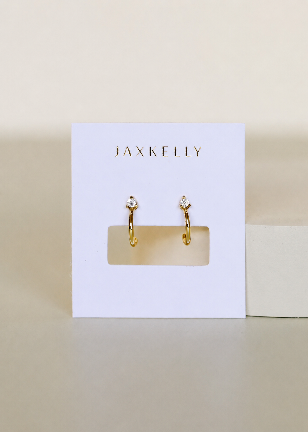 The JaxKelly Open Mini Hoop Earring - White is a pair of small gold huggie hoops with clear gemstone studs, shown on a white JaxKelly card against a neutral background.