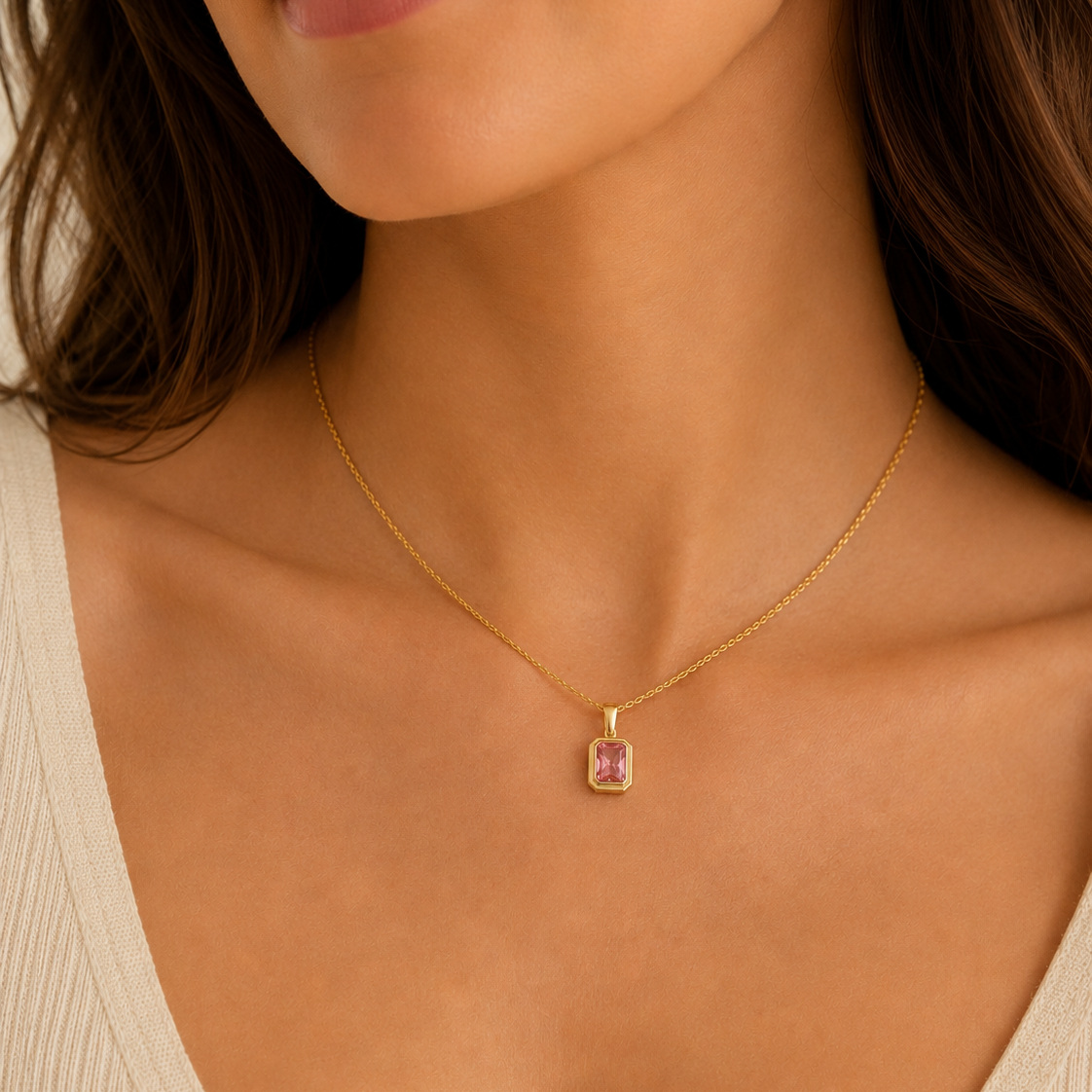 A woman with long brown hair, wearing a cream textured V-neck top, showcases the JaxKelly Rectangular Baguette Pendant Necklace in Light Pink, featuring an 18k gold plated bezel and pink baguette stone pendant.