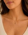 A woman with long brown hair, wearing a cream textured V-neck top, showcases the JaxKelly Rectangular Baguette Pendant Necklace in Light Pink, featuring an 18k gold plated bezel and pink baguette stone pendant.