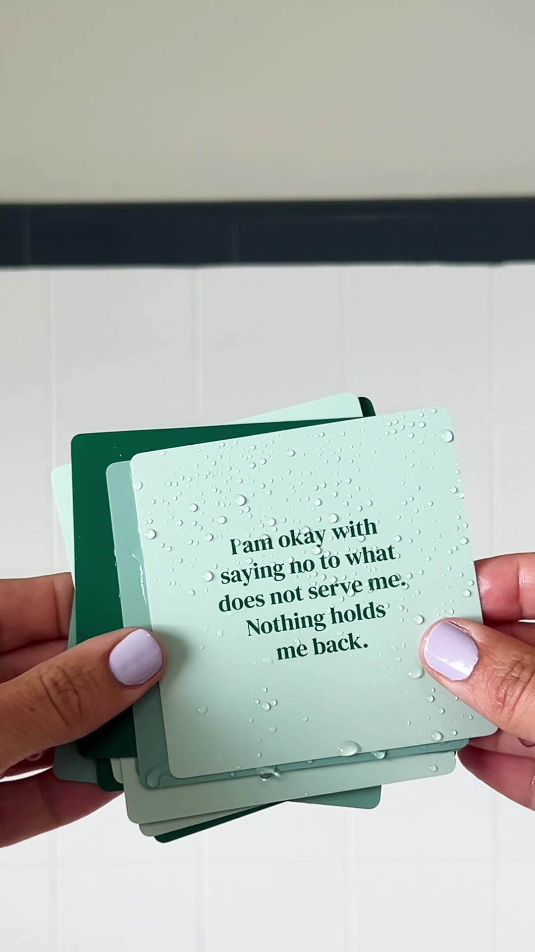JaxKelly's Abundance Shower Affirmations, slightly wet, being displayed and shuffled through by a pair of hands.