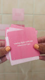 JaxKelly's Self-Love Shower Affirmations under running water, being placed on a tan-colored tile shower wall.