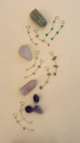 JaxKelly's Raw Gemstone Chain earrings displayed on a tan background, surrounded by crystals, and being picked up by a pair of hands one-by-one.