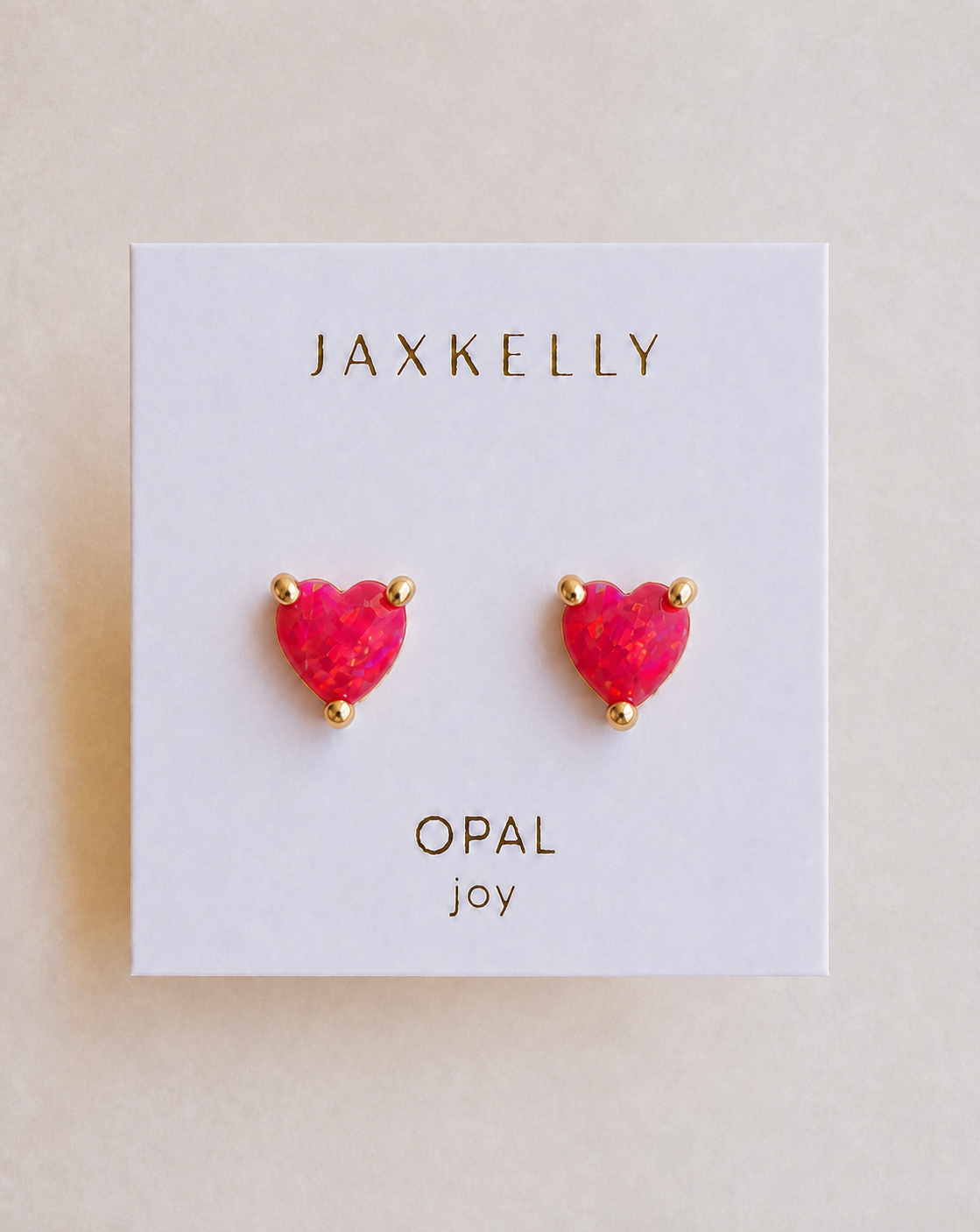 The JaxKelly Puffed Heart - Pink Opal stud earrings feature lab-grown pink opal and 18k gold plating, displayed on a white card that reads “JAXKELLY OPAL joy.”.