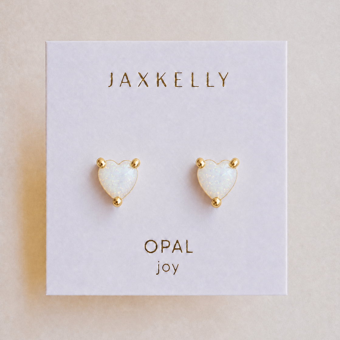 Elegant and hypoallergenic, the JaxKelly Puffed Heart - White Opal earrings feature heart-shaped opals with gold-plated studs, displayed on a white card labeled with “JaxKelly,” “Opal,” and “joy” in gold lettering—perfect for any occasion.