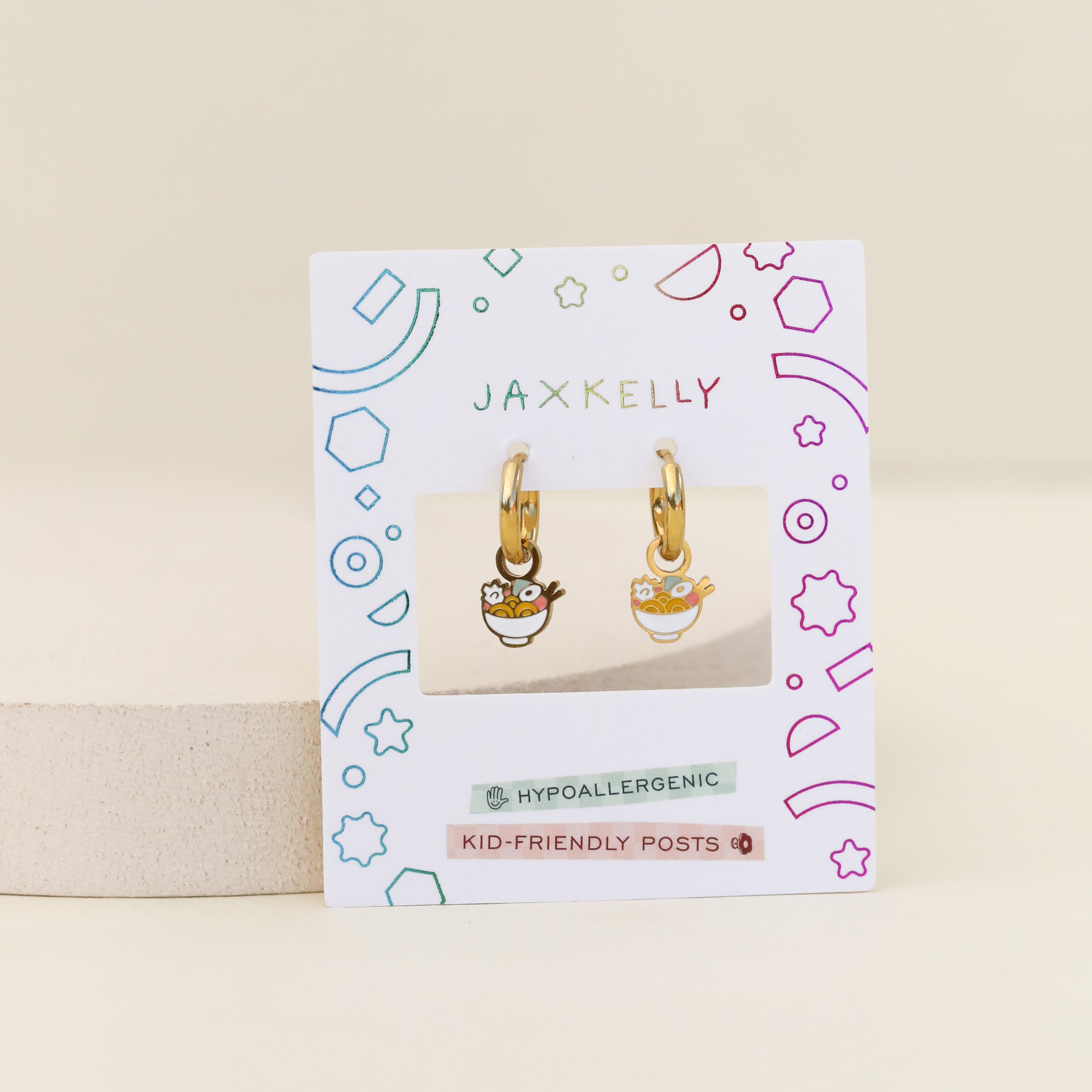 A pair of Ramen Enamel Charm Hoop kids earrings displayed on a JaxKelly card with colorful geometric designs labeled “hypoallergenic” and “kid-friendly posts.” Perfect for sensitive ears, these hoops feature fun ramen charms.