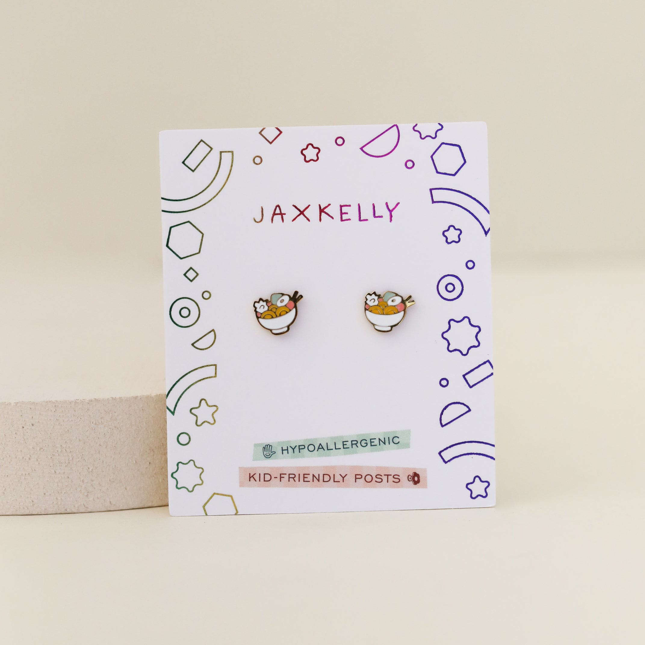 A pair of Ramen Enamel Flat Back Kids Earrings is displayed on a white JaxKelly card with colorful geometric doodles, noting the hypoallergenic earrings feature kid-friendly posts.