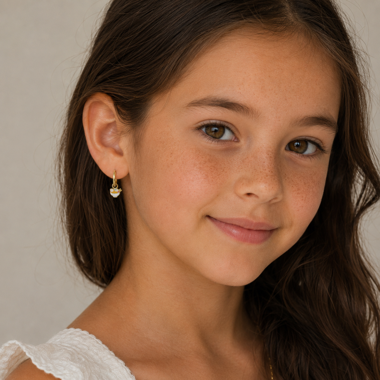 A young girl with long brown hair and brown eyes smiles softly, wearing a white top and JaxKelly's Ramen Enamel Charm Hoop earrings for sensitive ears, standing against a neutral background.