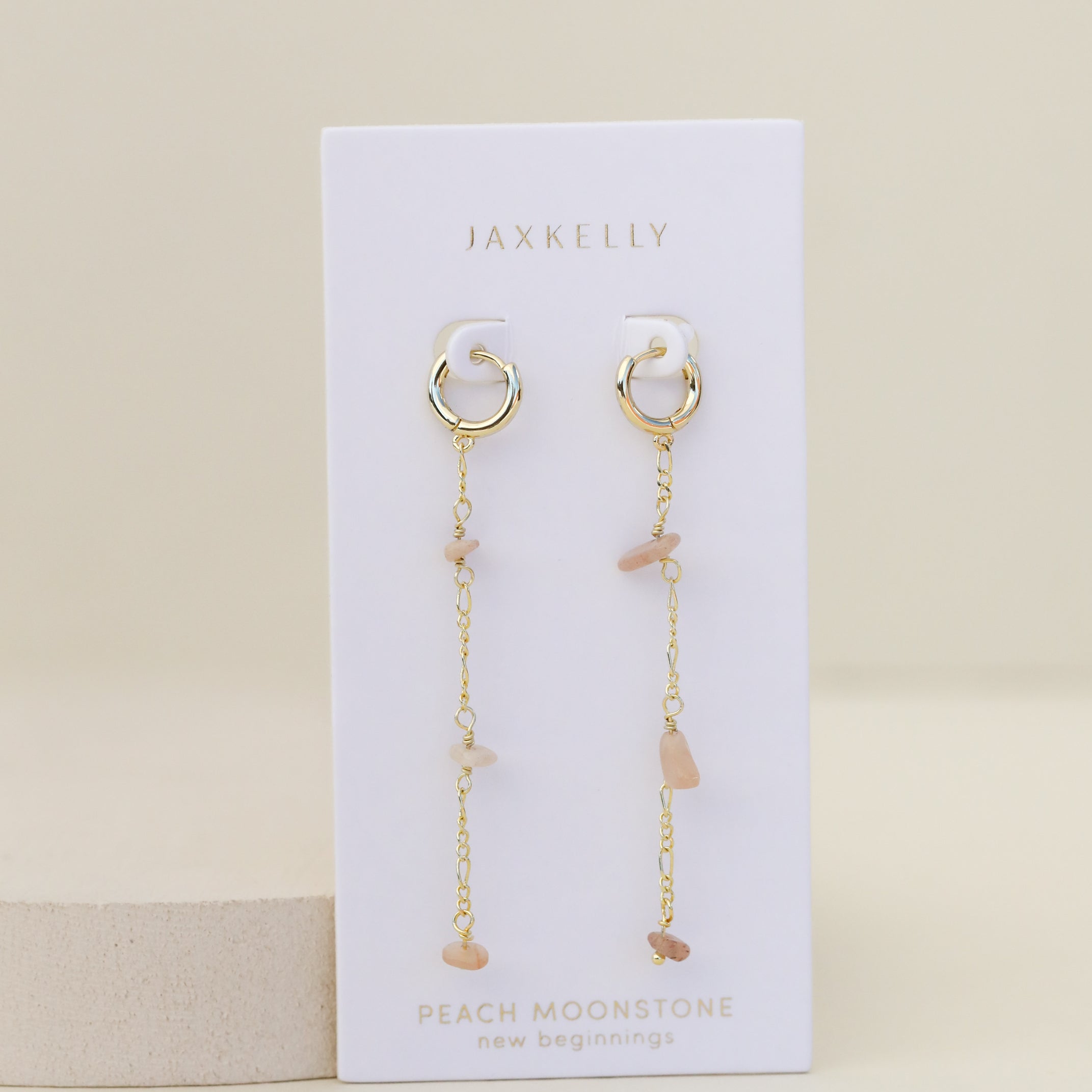 The Raw Gemstone Chain Drop Earring - Peach Moonstone features gold chain drop hoops with dangling chains and peach moonstone chips, displayed on a white JaxKelly card labeled "Peach Moonstone – new beginnings." Tarnish free for effortless elegance.