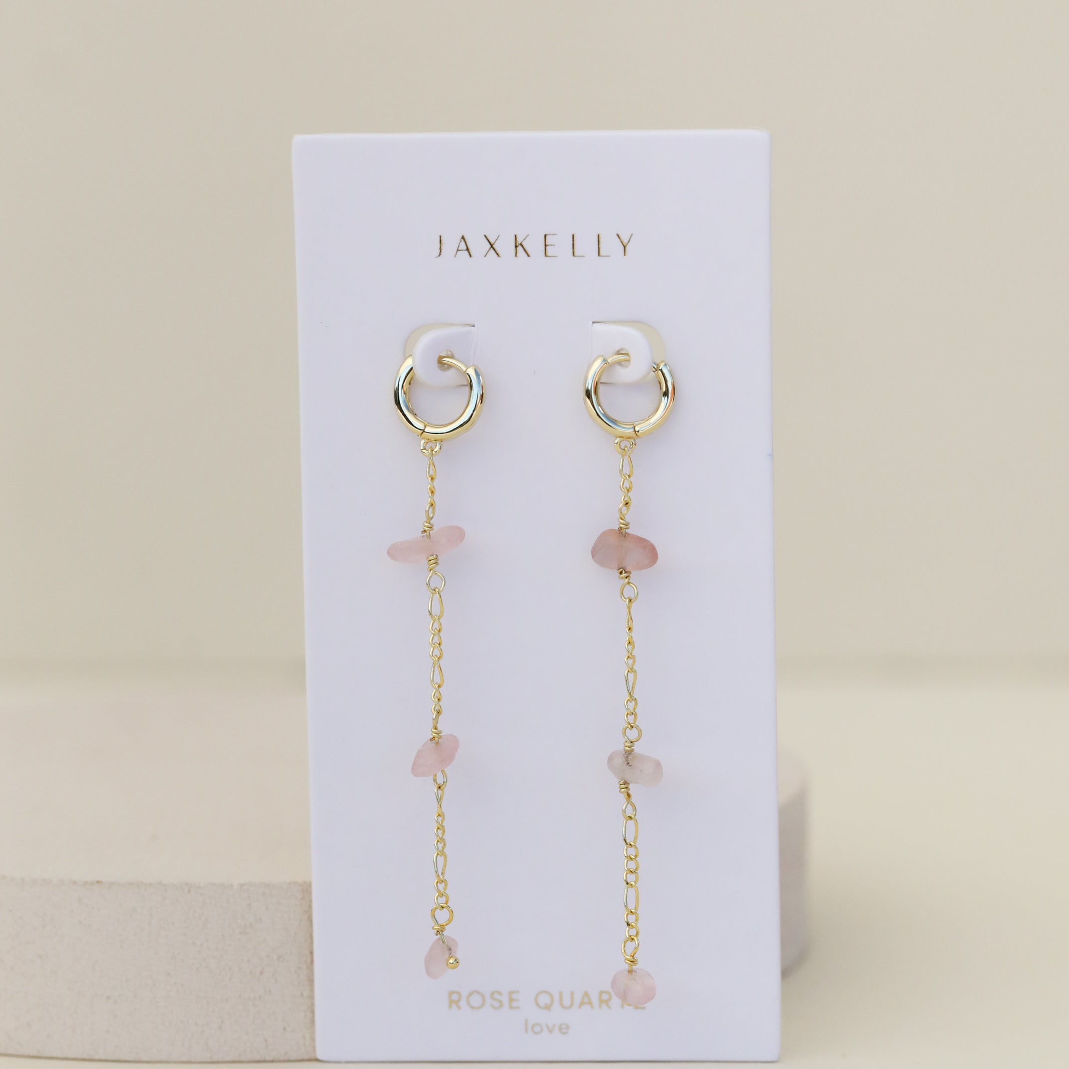 The Raw Gemstone Chain Drop Earring - Rose Quartz features gold chain drop hoops displayed on a white JaxKelly card labeled "Rose Quartz love," against a light beige background. These tarnish-free earrings add elegance to any look.