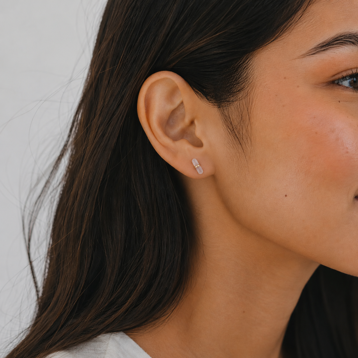 Close-up of a woman with long dark hair and smooth skin wearing JaxKelly's Rose Quartz Mineral Point - Love earring, a delicate rectangular gold stud. She’s in a white top against a light background—minimalist elegance.