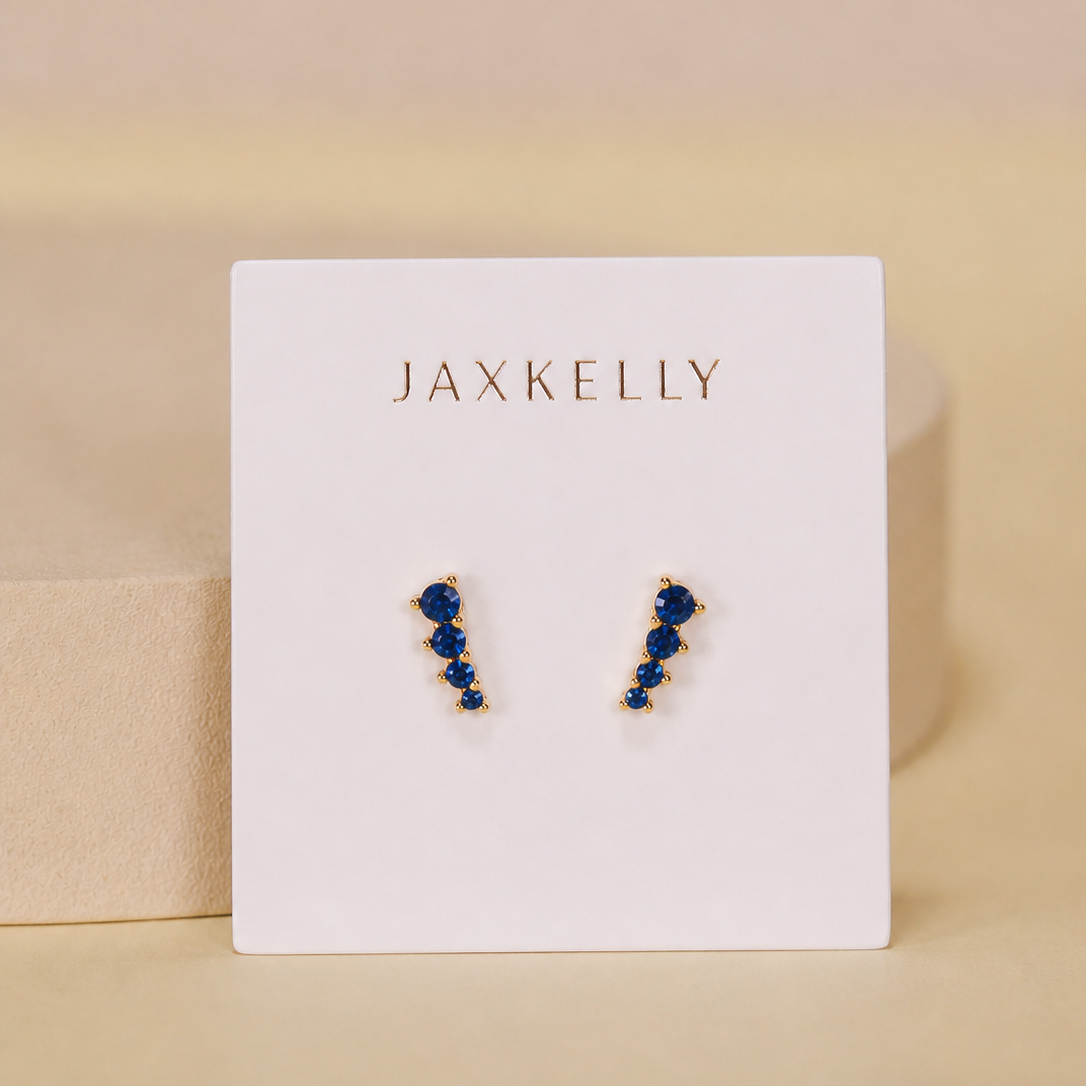 The Mini Crawler - Sapphire CZ - Gold Earrings by JaxKelly feature gold-plated studs with blue stones in a zigzag design. Displayed on a white JaxKelly card, these hypoallergenic earrings are perfect for sensitive ears.