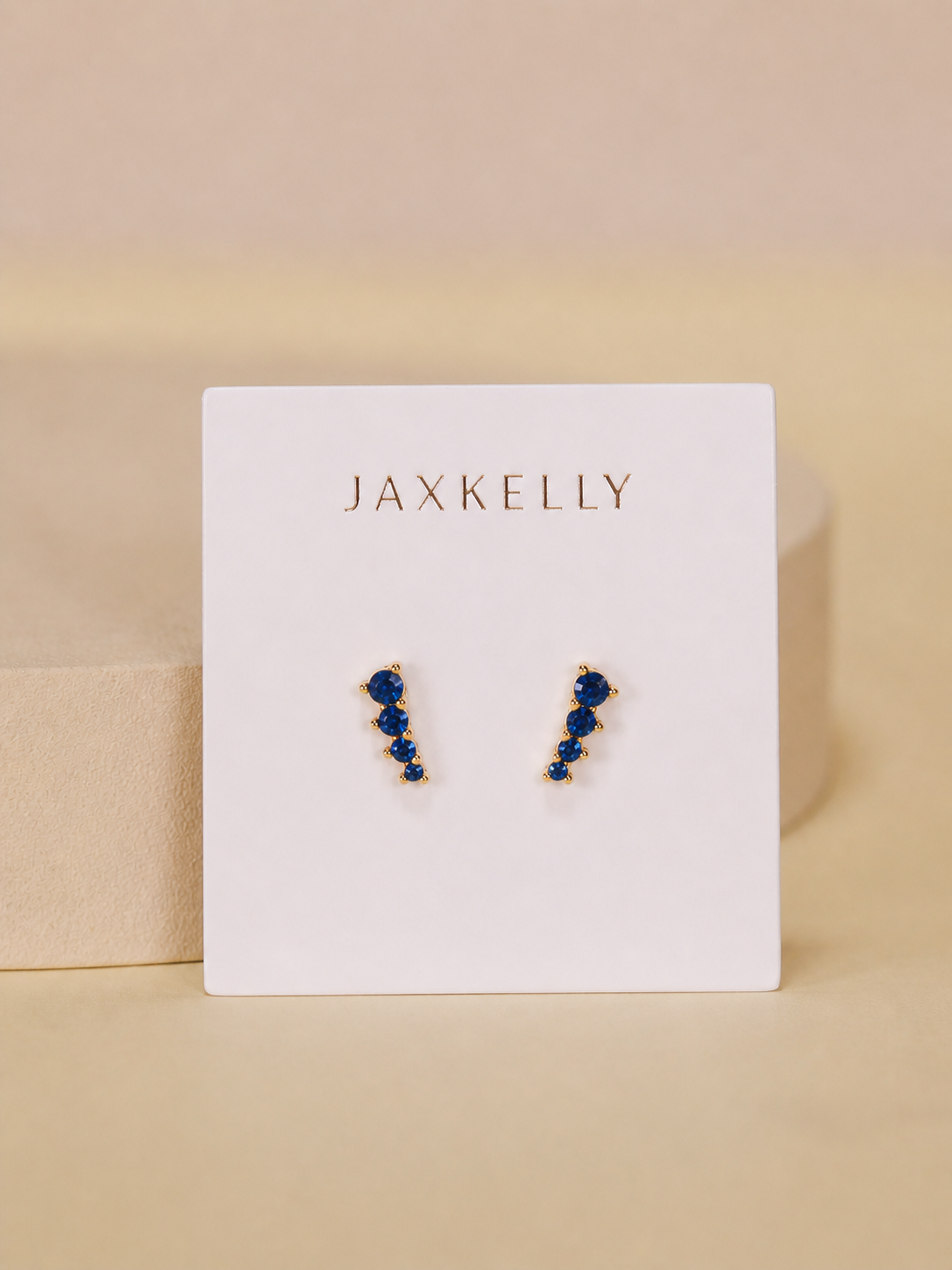 The Mini Crawler - Sapphire CZ - Gold Earrings by JaxKelly feature gold-plated studs with blue stones in a zigzag design. Displayed on a white JaxKelly card, these hypoallergenic earrings are perfect for sensitive ears.