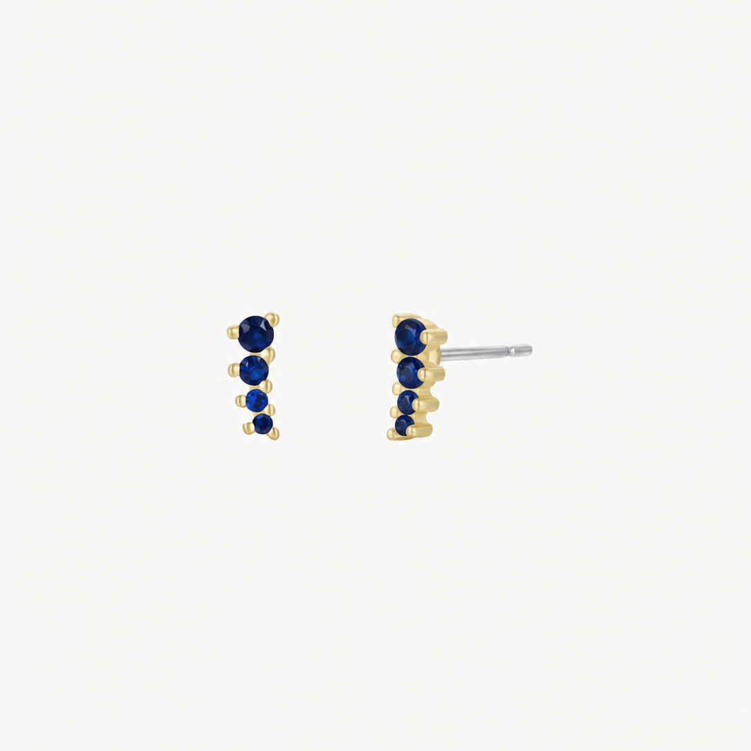 The Mini Crawler - Sapphire CZ - Gold Earrings by JaxKelly are small, gold-plated studs set with five dark blue sapphire CZ stones in a vertical line, offering hypoallergenic and elegant sparkle for any look.