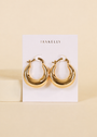 A pair of Sculptural Wide Hoop earrings in shiny 18K gold plating by JaxKelly are displayed on a white JaxKelly card against a beige background.
