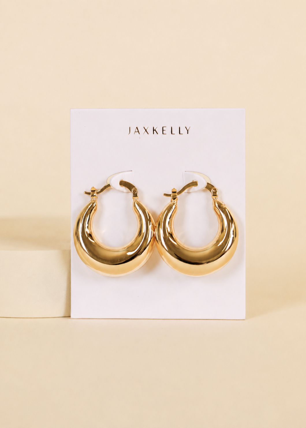 A pair of Sculptural Wide Hoop earrings in shiny 18K gold plating by JaxKelly are displayed on a white JaxKelly card against a beige background.