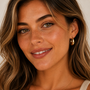 A woman with wavy brown hair, green eyes, and light freckles smiles softly. She wears JaxKelly’s tarnish-free Sculptural Wide Hoop earrings (18K gold plated) and a light top against a plain background.