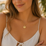 A woman outdoors smiles softly, wearing a cream sleeveless tie-front dress and the JaxKelly Seashell Pendant Necklace—an 18k gold plated piece with an adjustable chain.