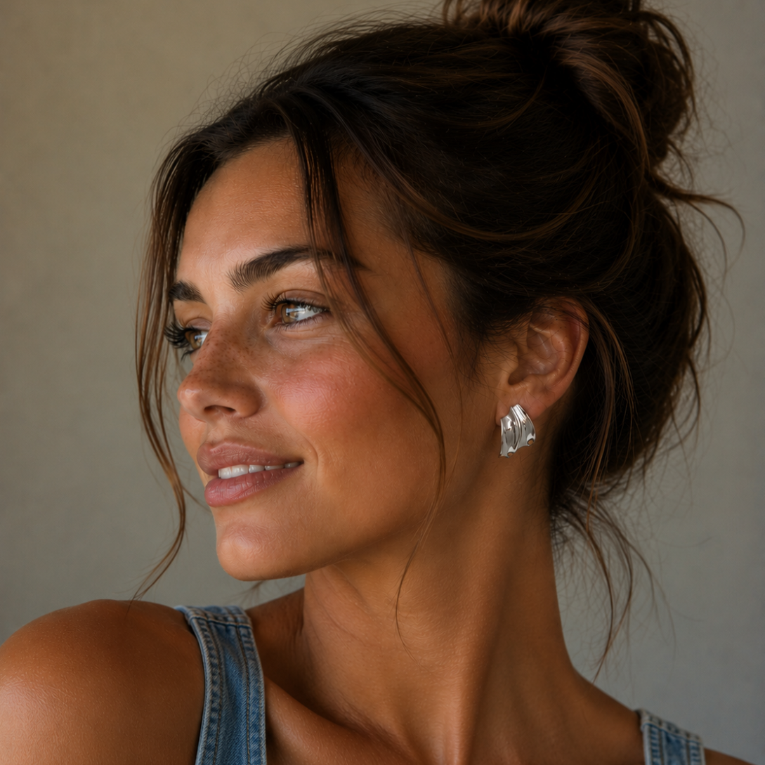 A woman with brown hair in a messy bun, wearing a sleeveless denim top and JaxKelly's Silver Bold Wave statement stud earrings, smiles softly as she looks to the side against a neutral background.