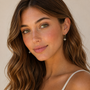 A woman with wavy brown hair and green eyes smiles gently, wearing a light tank top and the JaxKelly Starburst Mini Hoop earring, featuring a delicate starburst charm, posed against a neutral background.