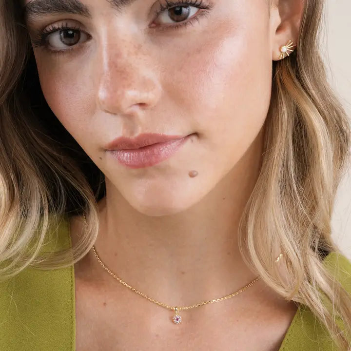 A woman with wavy blonde hair wears JaxKelly's Sun Ray - Mother of Pearl gold plated, leaf-shaped earrings with sterling silver posts and a delicate gold necklace. She has natural makeup and a light green top.