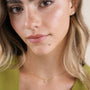 A woman with wavy blonde hair wears JaxKelly's Sun Ray - Mother of Pearl gold plated, leaf-shaped earrings with sterling silver posts and a delicate gold necklace. She has natural makeup and a light green top.