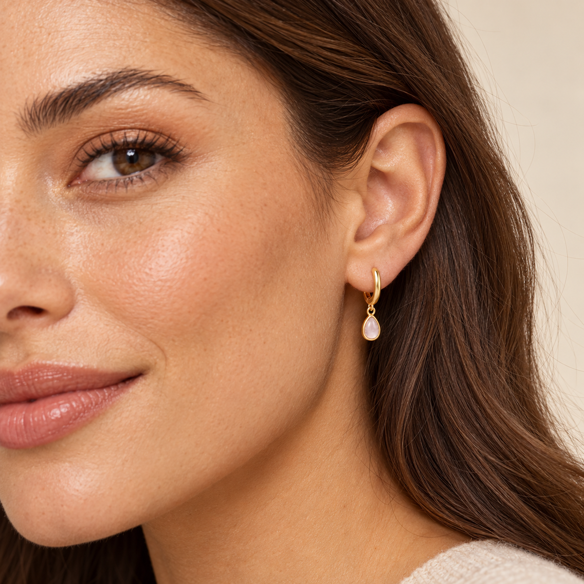 Close-up of a person wearing a JaxKelly Teardrop Charm Hoop - Rose Quartz, featuring a tarnish-free gold hoop earring with a dangling teardrop-shaped light pink stone charm.