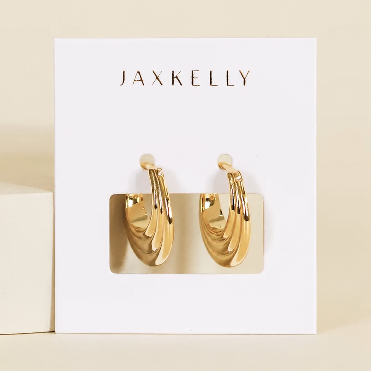 The JaxKelly Triple Arch Hoop Earrings feature a layered, twisted sculptural design in 18K gold, shown on a white branded card against a neutral background.