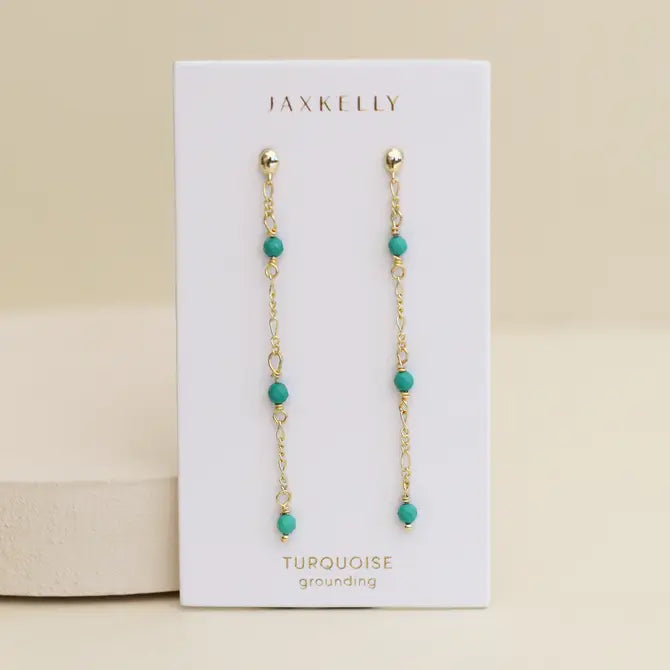 The JaxKelly Gemstone Chain Drop - Turquoise earrings, featuring small turquoise beads and 18k gold plating, are displayed on a white card labeled JaxKelly and Turquoise against a neutral background.
