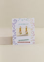 The Unicorn Enamel Charm Hoop features gold hoops with rainbow and cloud enamel charms, displayed on a JaxKelly card labeled hypoallergenic earrings and kid-friendly posts, surrounded by colorful geometric shapes.