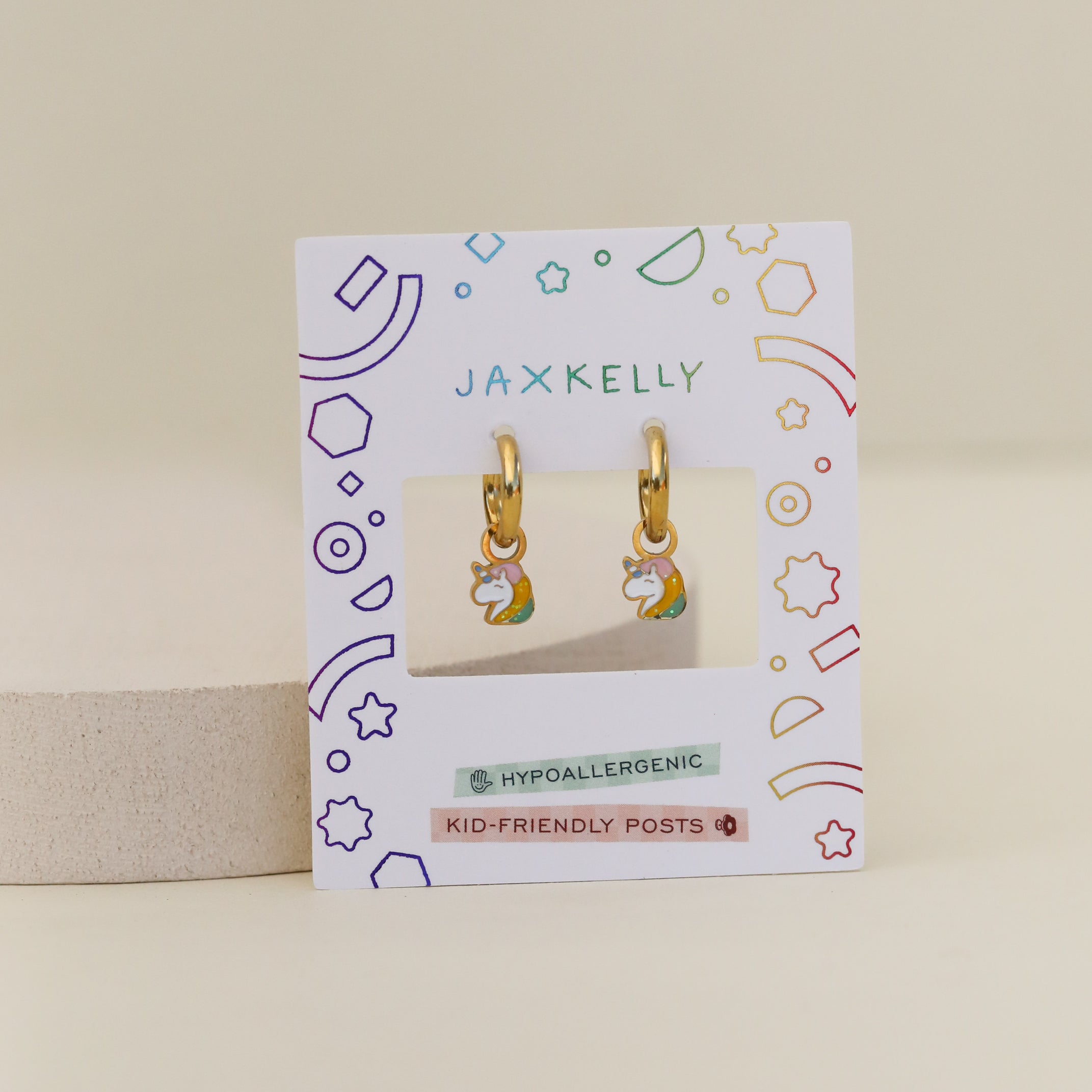 The Unicorn Enamel Charm Hoop features gold hoops with rainbow and cloud enamel charms, displayed on a JaxKelly card labeled hypoallergenic earrings and kid-friendly posts, surrounded by colorful geometric shapes.