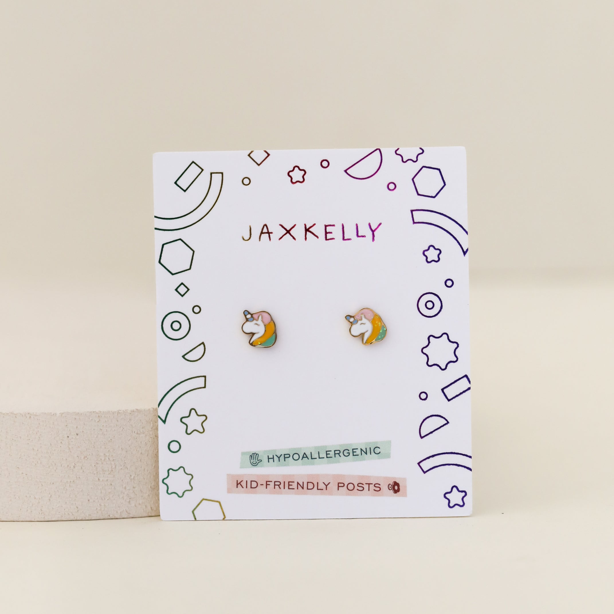 Unicorn Enamel Flat Back Kids Earrings with rainbow accents on a JaxKelly card with colorful geometric designs, labeled “hypoallergenic” and “kid-friendly posts.”.