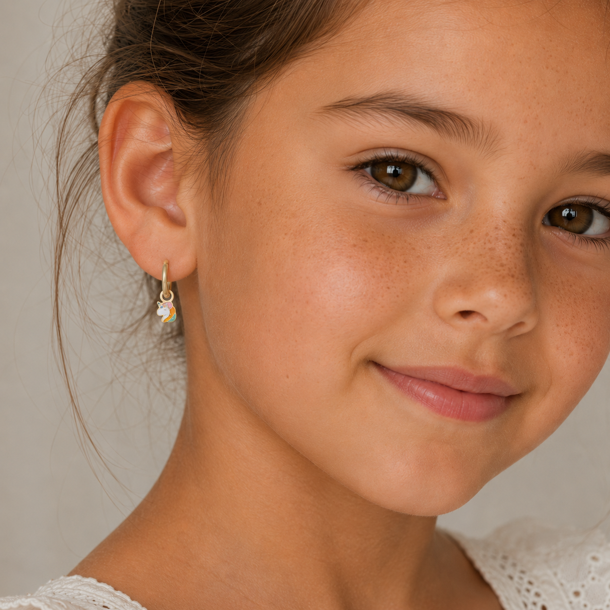 A young girl with brown eyes and freckled skin smiles softly, wearing the JaxKelly Unicorn Enamel Charm Hoop—ideal for kids and gentle on sensitive ears—with a white lace-detailed top against a neutral background.