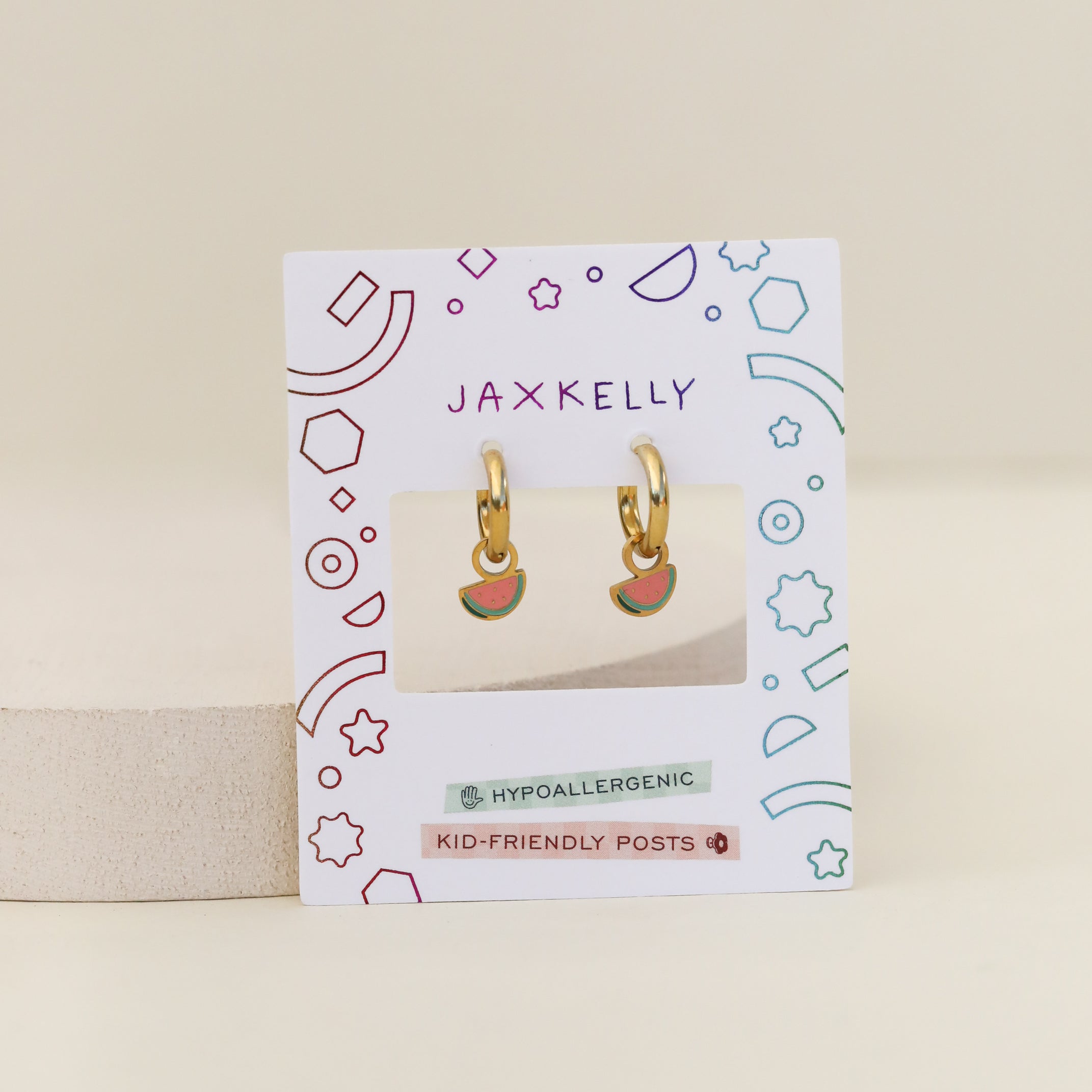 The Watermelon Enamel Charm Hoop earrings, ideal for sensitive ears, are shown on a JaxKelly card with colorful geometric patterns and labels stating Hypoallergenic and Kid-Friendly Posts.