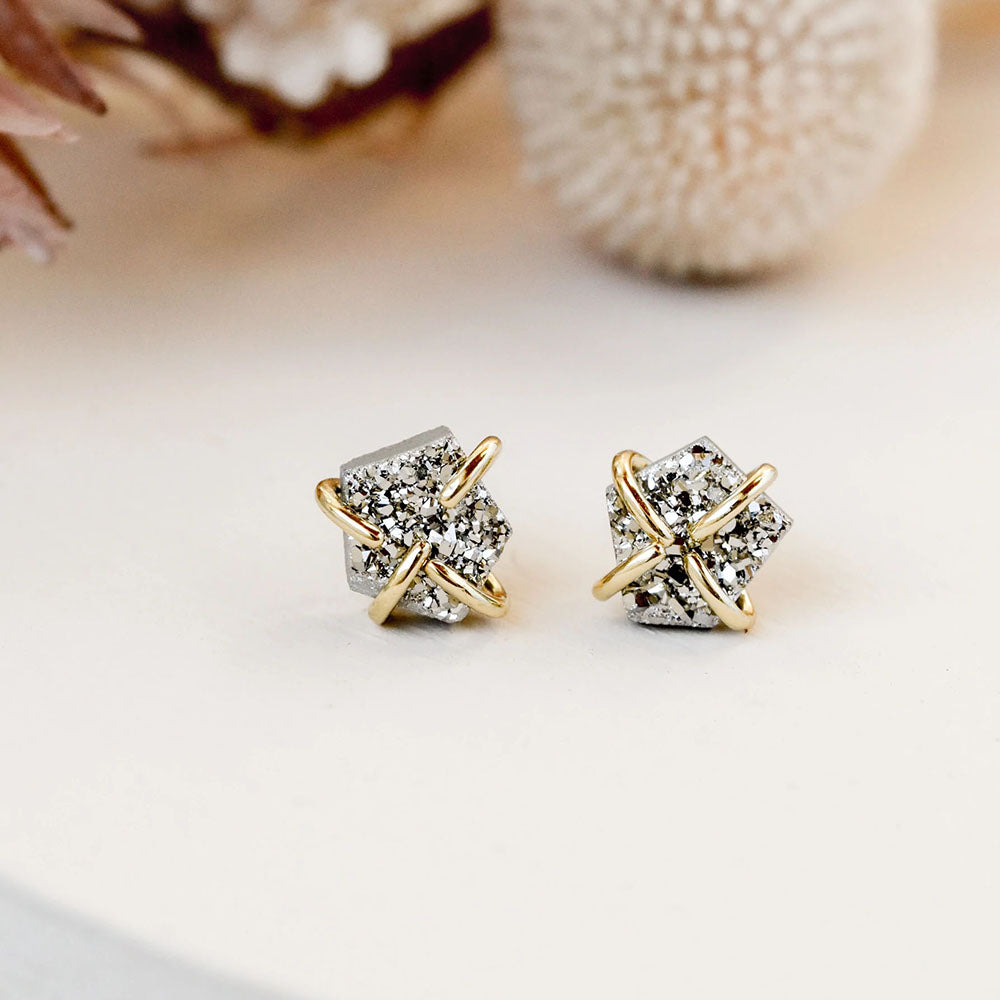 The Silver Druzy Prong earrings feature geometric studs with sparkling silver druzy stones wrapped in crisscrossed 18k gold plating, shown on a light surface with soft floral accents in the background.