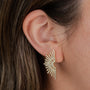 A woman's ear is shown wearing Sunburst Studs—gold vermeil earrings featuring thin, sparkling rays with small gems. Brown hair partially frames the ear.