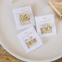 Three pairs of Pavé Gold Hoop 18k gold plated earrings on JAXKELLY cards are displayed on a round white dish set atop a light wooden surface.