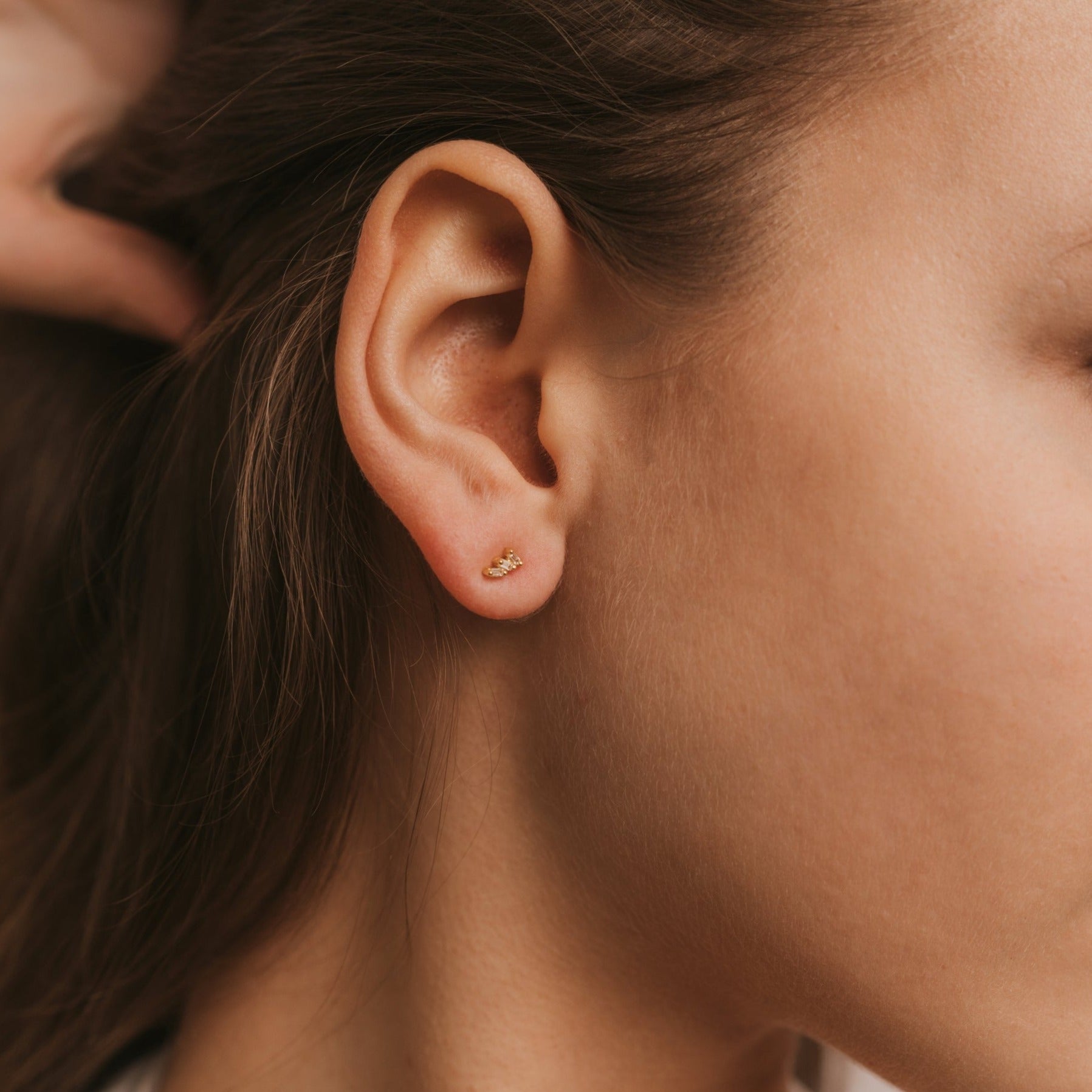 Close-up of a woman's ear adorned with the Offset Trio - White CZ stud earring, plated in 18k gold. Her brown hair is tucked behind her ear, drawing focus to the elegant earring and her natural skin texture against a soft background.