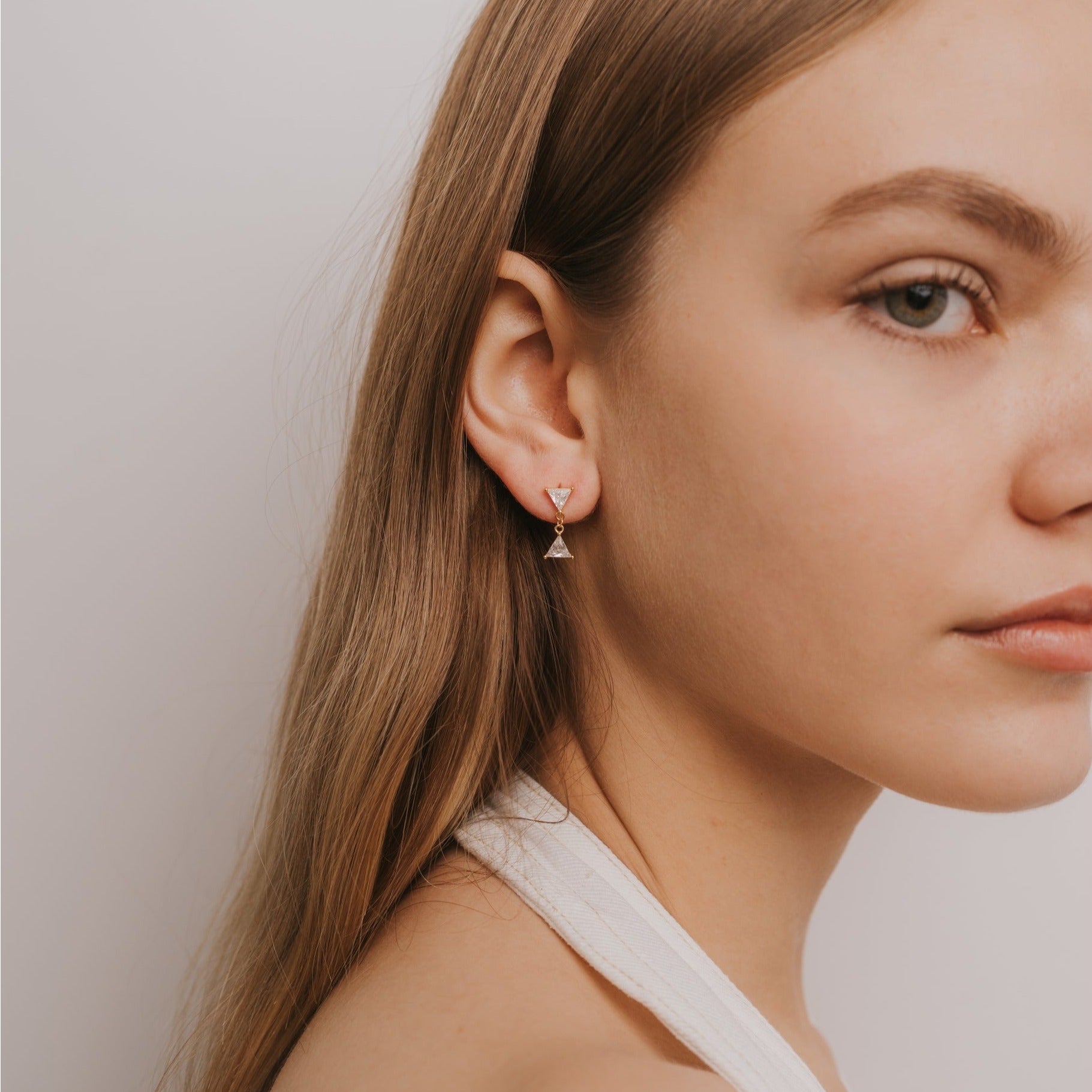 A young woman with long light brown hair wears a white halter top and the Dual Triangle Drop, a small, delicate CZ earring with sterling silver posts, while looking slightly away from the camera against a plain light background.
