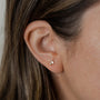Close-up of a person with straight brown hair wearing the Double Stud Stack: a small gold earring with two crossed rectangular gemstones—one a white baguette CZ—shining in 18k gold plating against a softly blurred background.