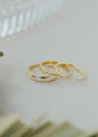 The Inset Baguette Ring, a gold stacking ring with small sparkling diamonds and sleek design, rests on a light surface. Background details include a blurred glass piece and green plant leaves. The ring features both rectangular and round stones.
