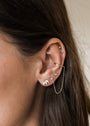 Close-up of a woman's ear with gold-plated piercings, including small studs, a hoop with a delicate chain, and the White Opal Crown Stud; strands of brown hair frame the ear.
