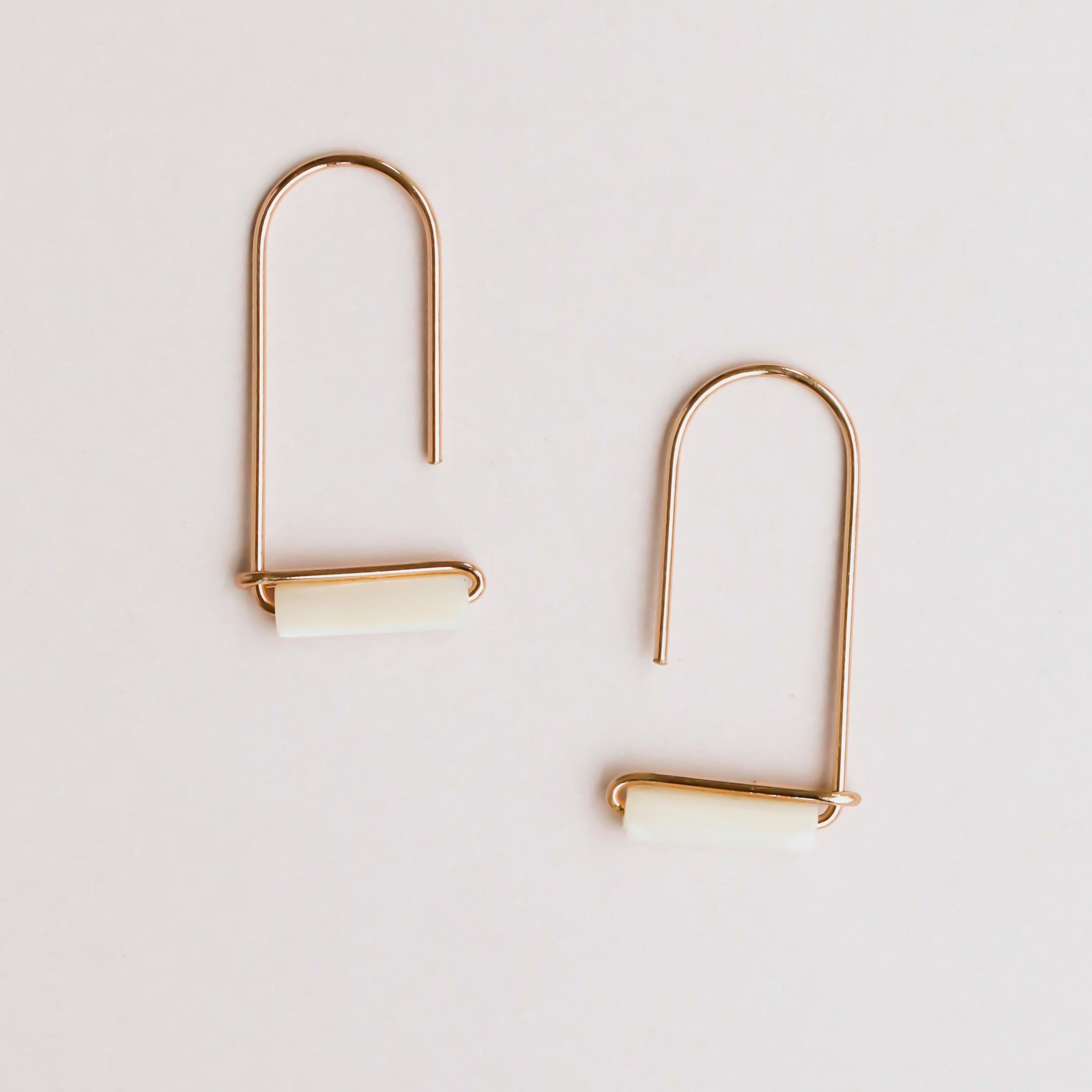 The Mother of Pearl Gemstone Drop - Calming earrings feature minimalist gold-plated wire, U-shaped hooks, and horizontal white Mother of Pearl cylinder accents, displayed on a simple light background.